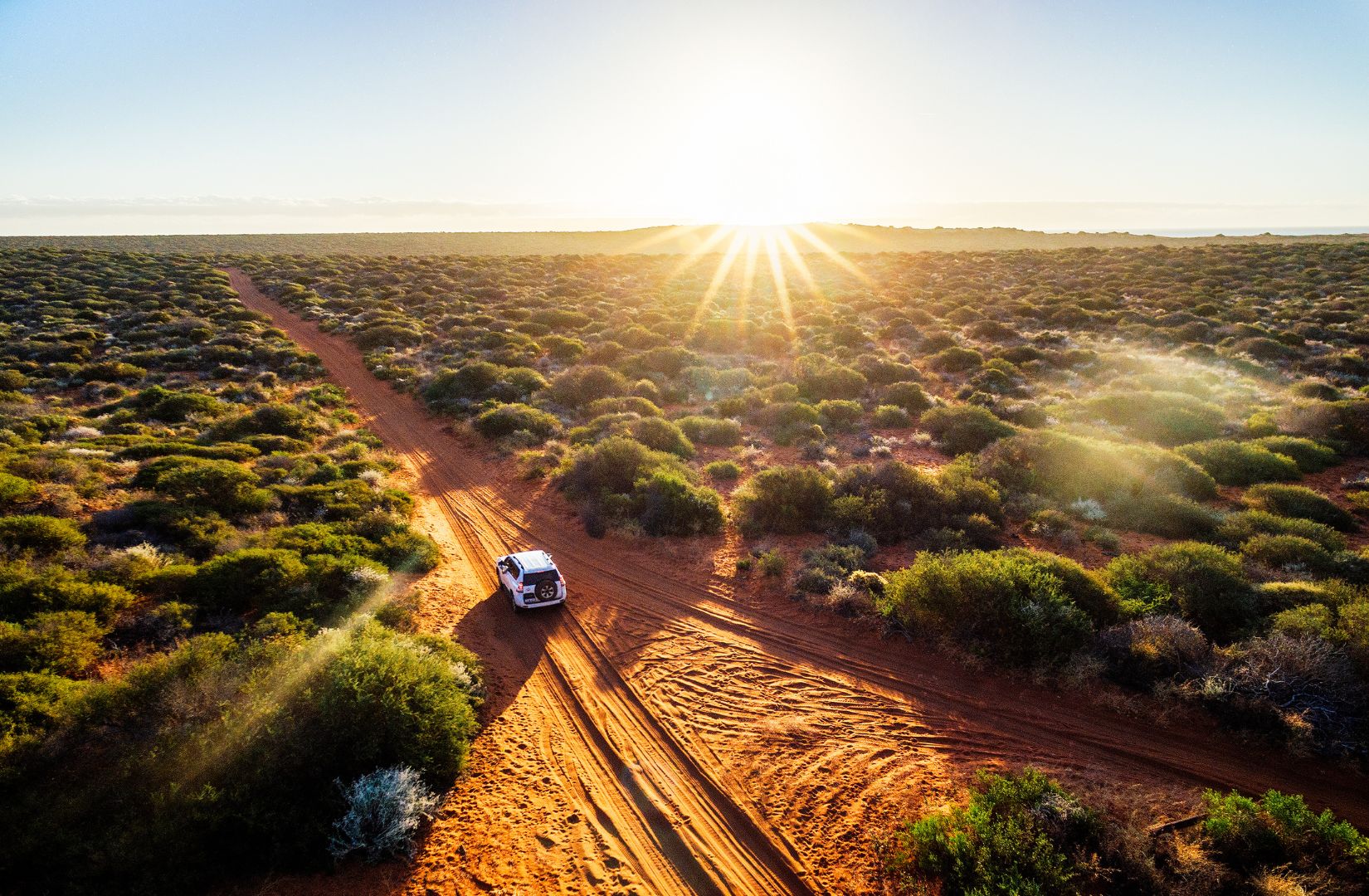 A car travels down a red dirt track in remote Australia. The sun is setting in the background of the image.